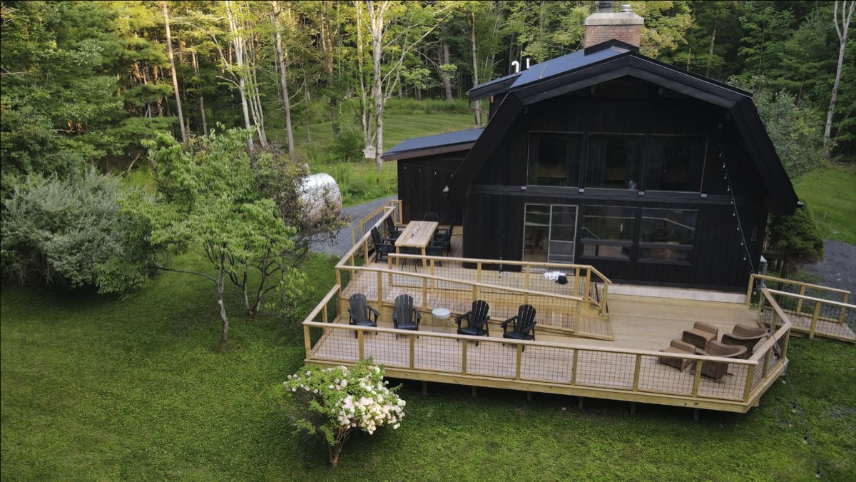 The Hideaway&rsquo;s front deck with Adirondack chairs, an outdoor dining table, and the barrel sauna at the tree line